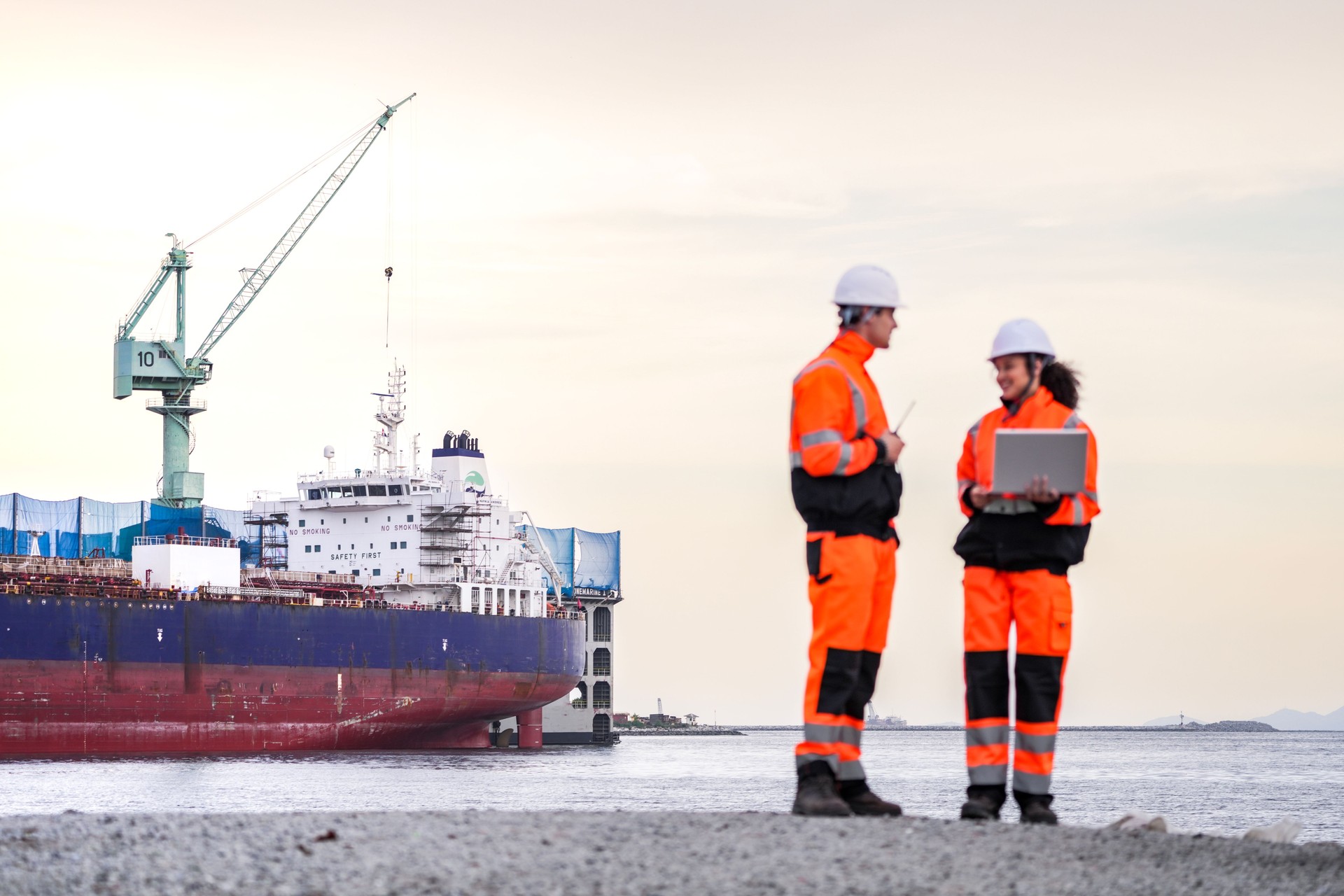 Two engineers wearing high-visibility safety gear and hard hats review plans on a laptop at a shipyard, with a large cargo ship and cranes in the background during sunset.
