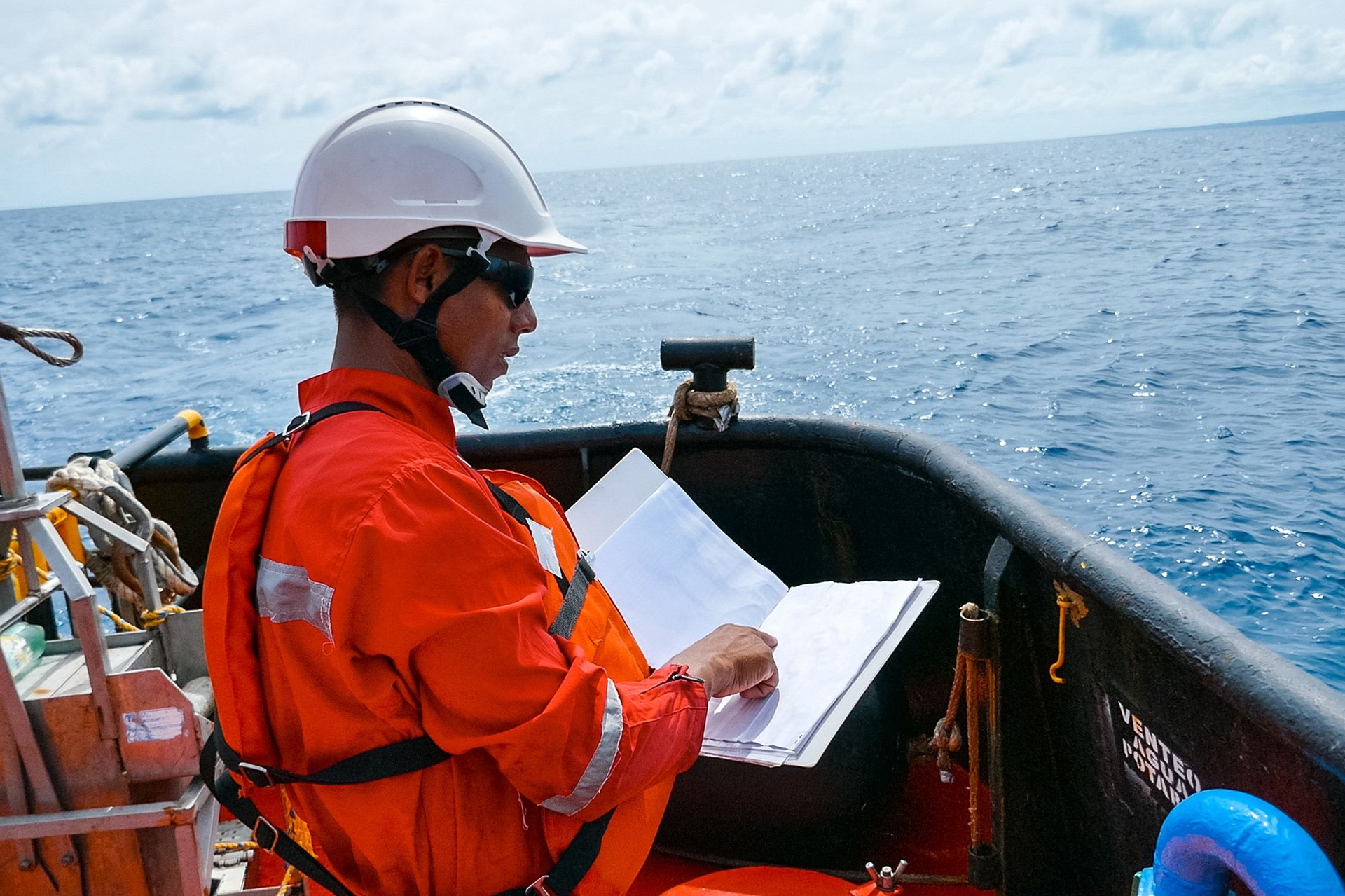 Safety officer, standing on an industrial ship, wearing overalls, a helmet, safety goggles and holding a clipboard with checklists.